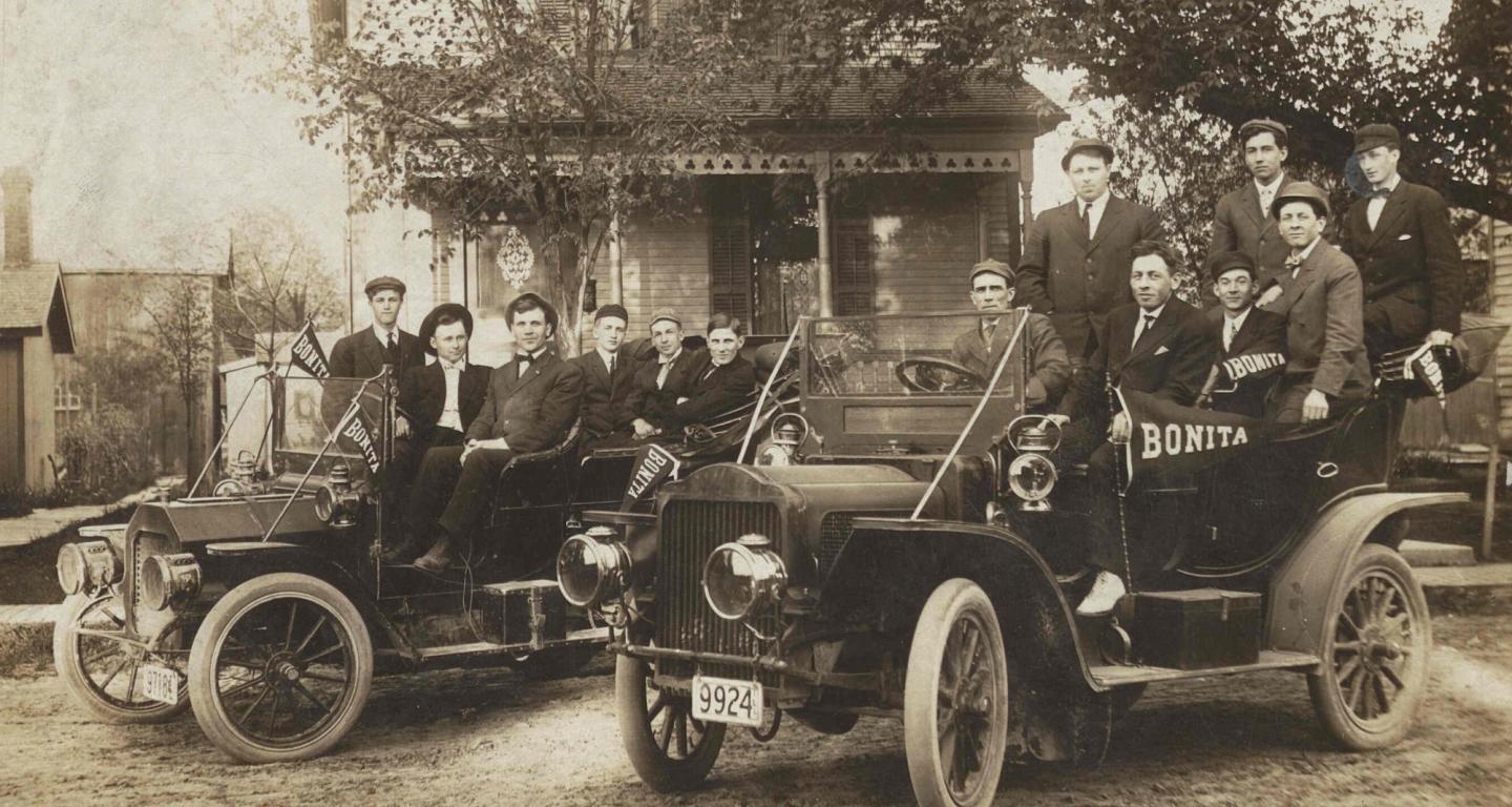 Several young people sit in two 1930s automobiles in front of a house